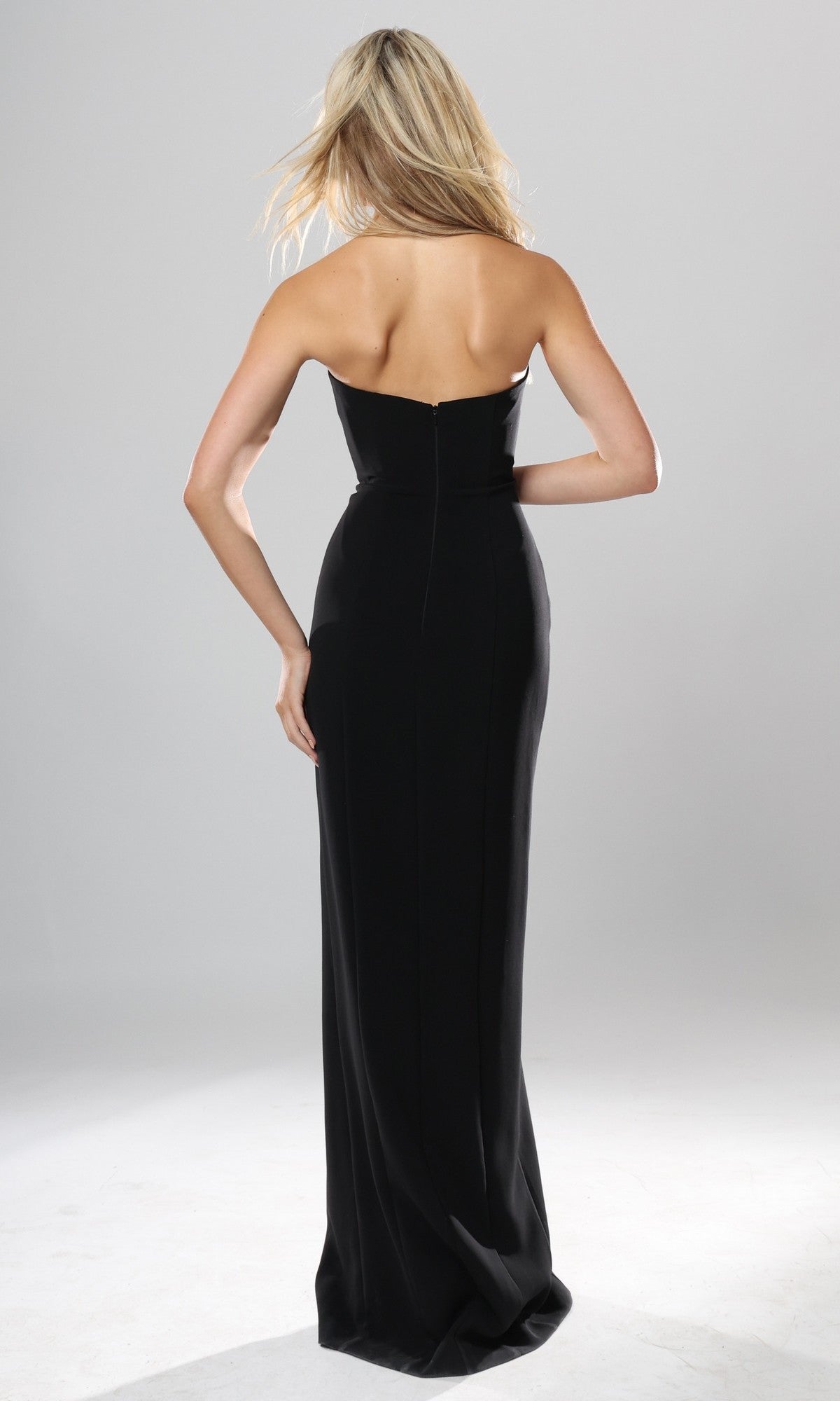 A woman with long blonde hair is wearing the Jovani 53108 Long Formal Dress, a strapless, floor-length black gown, standing with her back to the camera against a plain light gray background.